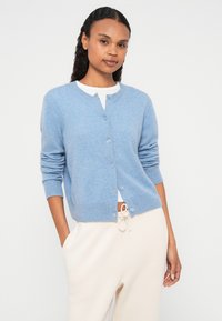 Svetlomodrý kardigán z jemného materiálu, s okrúhlym výstrihom, zapínaním na gombíky a rebrovanými manžetami. Nosený cez biele tričko.