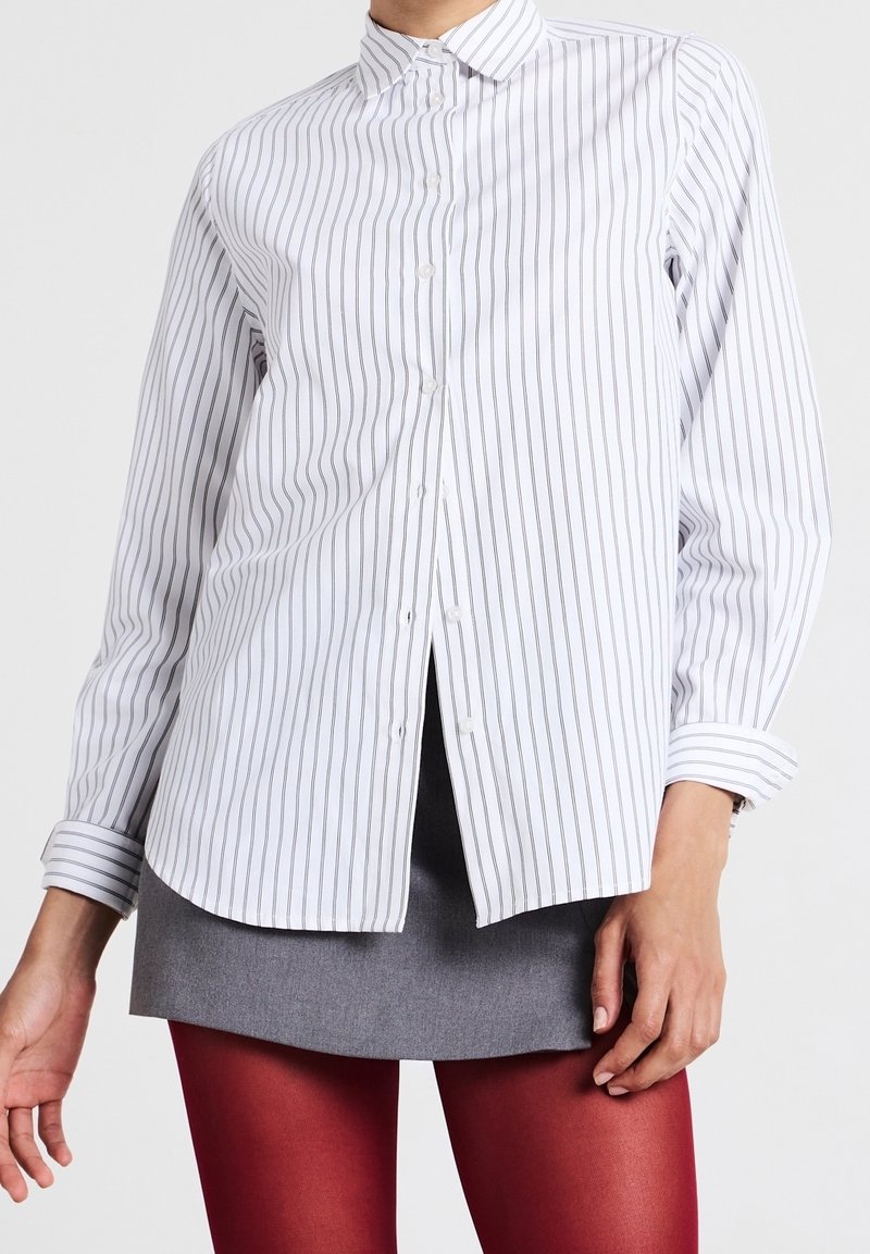 Personne portant une chemise blanche à manches longues avec de fines rayures noires verticales, une jupe grise et des collants rouges, sur un fond uni.