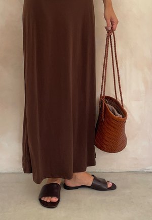 Personne portant une longue robe marron, des sandales ouvertes marron foncé, tenant un sac à main marron tressé contre un mur de fond neutre.