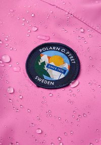 Vaaleanpunainen vedenpitävä kangas, jossa on pyöreä brodeerattu logo, jossa on vuoria ja aurinko, sekä teksti "POLARN O.PYRET SWEDEN" ja vesipisaroita.