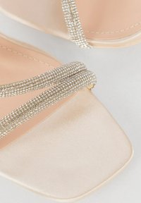 Beige Satin-Slipper-Sandalen mit quadratischer Zehenform, ausgestattet mit zwei glitzernden Riemen und einer glatten, glänzenden Oberfläche.