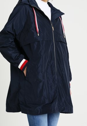 Laivastonsininen hupparillinen takki, jossa edessä vetoketju, punavalkoraidalliset kiristysnauhat ja vastaavat raidalliset hihansuut, päällä sinisten farkkujen kanssa.