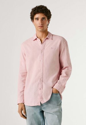 Jeune homme aux cheveux bouclés portant une chemise rose claire à boutons et un jean bleu clair, debout avec une main dans la poche devant un fond uni.