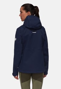 Navy blue waterproof jacket with a hood, featuring a logo on the back, side pockets, and adjustable cuffs. Smooth, lightweight fabric.