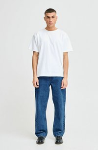 Camiseta blanca de algodón de manga corta combinada con unos pantalones vaqueros oscuros de corte holgado y zapatos negros, de pie frente a un fondo neutro.
