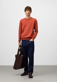 Giovane uomo che indossa un maglione color ruggine, jeans blu scuro, scarpe di pelle marrone, mentre tiene una grande borsa tote di pelle marrone scuro, in piedi all'interno.