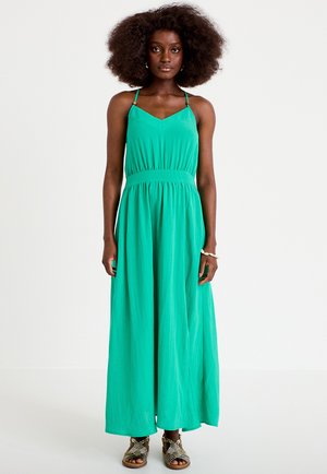 Kvinde stående iført en ærmeløs grøn maxikjole med tynde stropper og flade vævede sandaler mod en ensfarvet hvid baggrund.