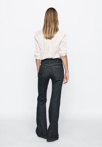 Fitted dark denim jeans with flared legs, back pockets, and a black waistband detail, paired with a wrinkled off-white shirt.
