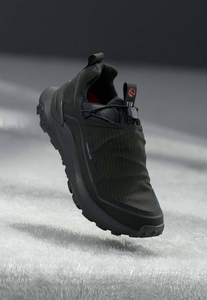 Baskets Timberland noires avec semelle texturée et languettes, positionnées au-dessus d'une surface douce et claire avec un arrière-plan ombragé.