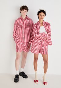 Rot-weiß karierte Hemden und Shorts mit Knopfdetails und Taschen. Das Modell trägt schwarze Schuhe, während eine Frau rote High Heels mit Socken trägt.