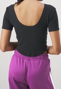 Czarny krótki top z rękawem, z tyłem w kształcie litery U, o dopasowanym kroju. Noszony z fioletowymi spodniami dresowymi, które mają elastyczny pas i boczne akcenty.