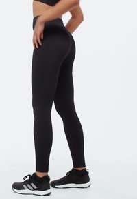 Leggings noirs taille haute en tissu extensible, présentant un design ajusté et une texture lisse, assortis à des chaussures de sport noires.