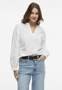 Hvid langærmet bluse med V-hals og puffede ærmer, parret med højere talje blå denim jeans og et brunt bælte med en sølvspænde.