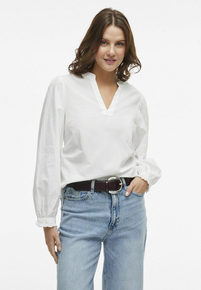 Hvid langærmet bluse med V-hals og puffede ærmer, parret med højere talje blå denim jeans og et brunt bælte med en sølvspænde.