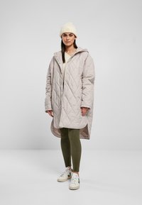 Paksu vaalea beige tikattu takki hupulla, sivutaskuilla ja napilla; päällä kreeminvärinen toppi ja oliivinvihreät legginsit, yhdistetty valkoisiin lenkkikenkiin.