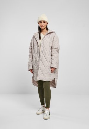 Leicht puffige, hellbeige gesteppte Jacke mit Kapuze, seitlichen Taschen und Druckknöpfen; getragen über einem cremefarbenen Oberteil und olivgrünen Leggings, kombiniert mit weißen Sneakers.