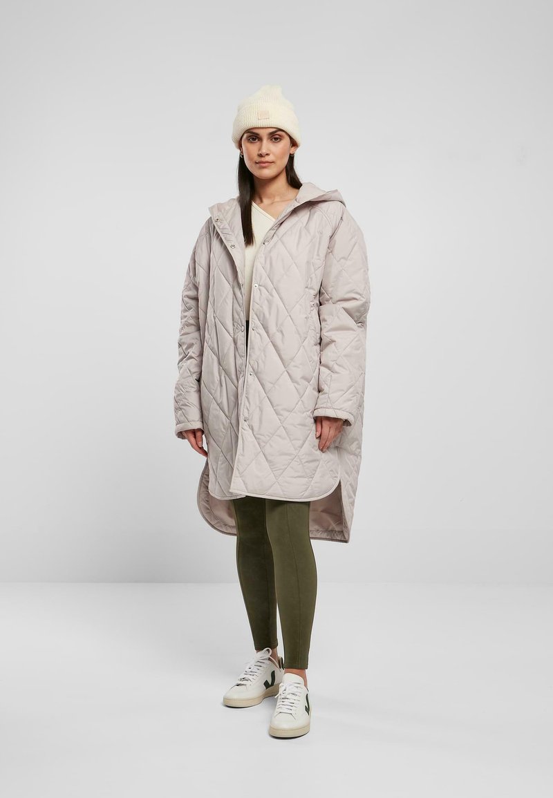 Dikke lichtbeige gewatteerde jas met capuchon, zijzakken en drukknoopsluiting; gedragen over een crèmekleurige top en olijfgroene leggings, gecombineerd met witte sneakers.