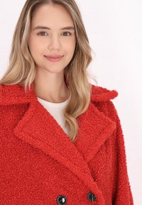 Cappotto in pelliccia sintetica rossa con design doppiopetto, ampio colletto e bottoni neri. Tessuto morbido e maniche lunghe, adatto per l'inverno.