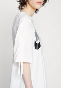 Camiseta blanca de gran tamaño hecha de algodón, con un logo negro de Nike. El diseño incluye lazos en los laterales de las mangas y un corte relajado.