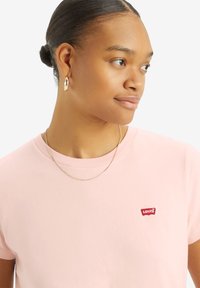 T-shirt in cotone rosa chiaro con un patch del logo rosso Levi's sul lato sinistro del petto. Texture liscia, scollo rotondo classico e maniche corte.