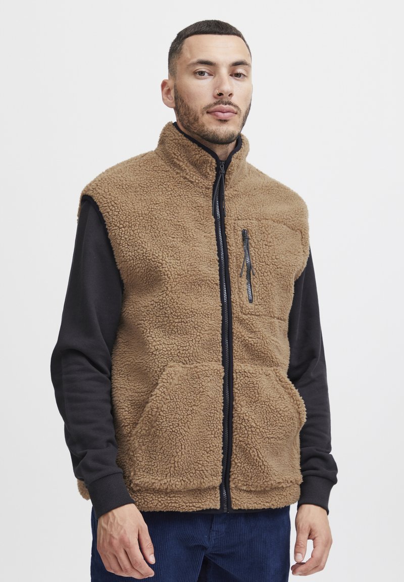 Vest din fleece maro cu guler înalt, cu fermoar pe față și două buzunare mari în față. Purtați peste un tricou negru cu mânecă lungă și mansete cu nervuri.
