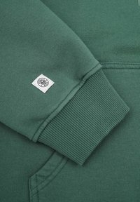 Mörkgrön sweatshirtmaterial med ribbad mudd, med en liten vit etikett med svart text och logotyp på vänster ärm.