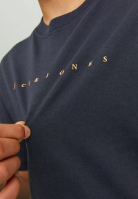 Tamsiai mėlyna medvilninė marškinėliai su apvaliame kakle, su logotipu, atspausdintu vario spalvos šriftu ant krūtinės. Lygi tekstūra, prigludusi forma.