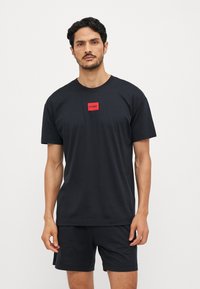 T-shirt en coton noir avec un logo carré rouge portant le texte "HUGO". Design simple avec col rond, manches courtes, porté avec un short assorti.
