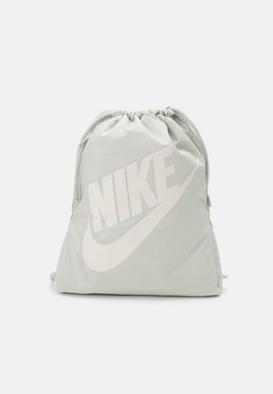 Hellgrauer Kordelzugbeutel mit großem weißem Nike-Logo und Swoosh auf der Vorderseite vor einfachem weißem Hintergrund.