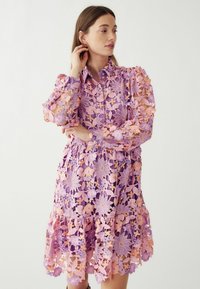 Nėriniuotas suknelė levandų ir švelnios rožinės spalvos gėlių raštu, su apykakle, ilgomis rankovėmis ir platėjančiu sijonu. Lengva ir tekstūruota.