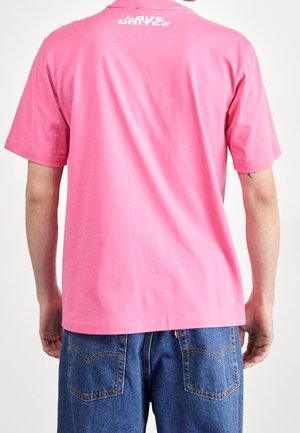 Mann trägt ein schlichtes pinkes T-Shirt mit weißem Text nahe dem Kragen und blaue Jeans, gezeigt von hinten vor einfarbigem Hintergrund.