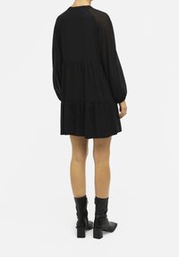 Robe noire avec des manches longues en tulle et une jupe à volants. Le design présente une coupe décontractée et est assorti à des bottines noires.