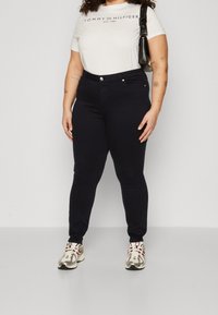 Persona vestida con una camiseta blanca de Tommy Hilfiger, pantalones negros de tiro alto, zapatillas blancas, llevando un bolso negro, de pie contra una pared blanca.