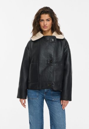 Un modèle portant une veste en cuir noire avec un col en peau lainée couleur crème et un jean bleu, debout devant un fond blanc uni.