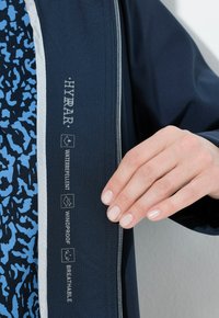Giacca navy con un esterno testurizzato. Presenta il brand HYRAR e icone per caratteristiche idrorepellenti, antivento e traspiranti. Chiusura con zip argentata.