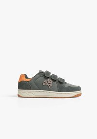 Zapatilla baja gris con tres tiras de velcro, acento naranja en el talón, suela beige y parche de calavera y huesos cruzados en el lateral.