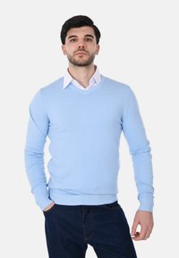 Uomo con capelli scuri e barba che indossa un maglione a V blu chiaro sopra una camicia bianca con colletto e jeans blu scuro su uno sfondo semplice.