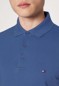 Blå polotröja med en texturerad tyg, tre knappar vid kragen och en liten broderad logotyp i röd, vit och blå på bröstet.