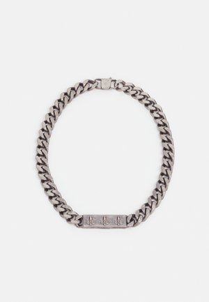 Bracelet chaîne épaisse couleur argent avec une plaque rectangulaire ornée de trois symboles fleur-de-lis gravés et une fermeture à boîte.