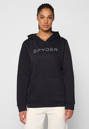 Frau mit geflochtenem Haar, die einen schwarzen Kapuzenpullover mit "SPYDER"-Aufschrift und weiße Hosen trägt, steht vor einem schlichten, hellen Hintergrund.