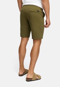 Hombre con pantalones cortos verde olivo hasta las rodillas y sandalias de ante color beige, de pie contra un fondo blanco liso.