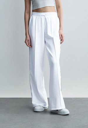 Osoba oblečená v bielych nohaviciach so širokými nohavicami s bočnými pruhmi, biele tenisky a biely crop top, stojaca na sivom podlaží pred jednoduchou stenou.