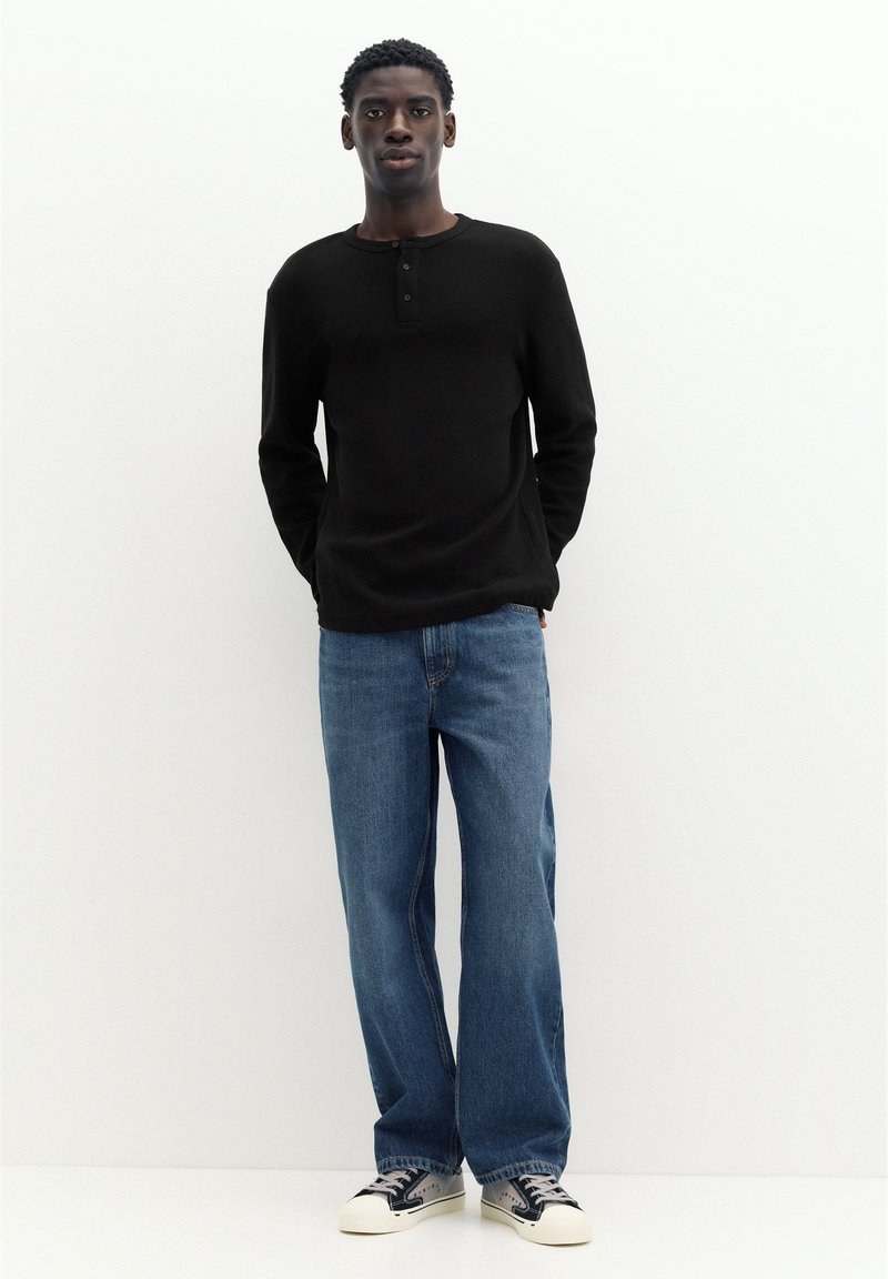 Jeune homme debout, les mains dans le dos, portant une chemise noire à manches longues, un jean bleu et des baskets noires et blanches sur fond blanc.