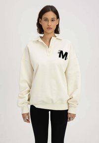 Crème-kleurige pullovertrui met een kraag, knopen aan de voorkant en een zwart geborduurd "M" logo. Lange mouwen en geribbelde zoomdetails.