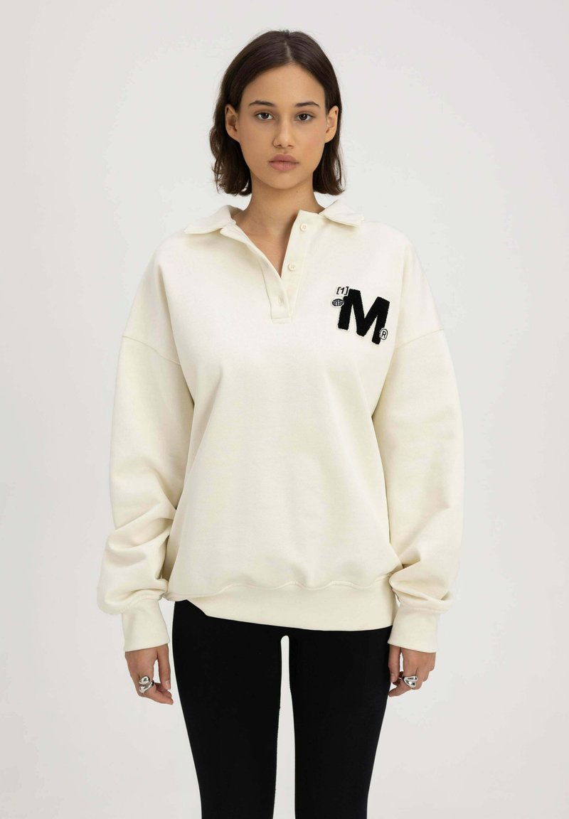 Crème-kleurige pullovertrui met een kraag, knopen aan de voorkant en een zwart geborduurd "M" logo. Lange mouwen en geribbelde zoomdetails.