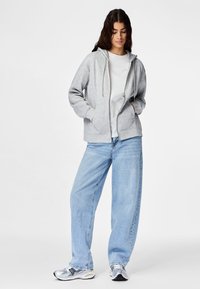 Mujer con cabello largo y oscuro, vistiendo una sudadera con capucha y cremallera de color gris claro, camiseta blanca, vaqueros sueltos azul claro y zapatillas plateadas, de pie con las manos en los bolsillos.