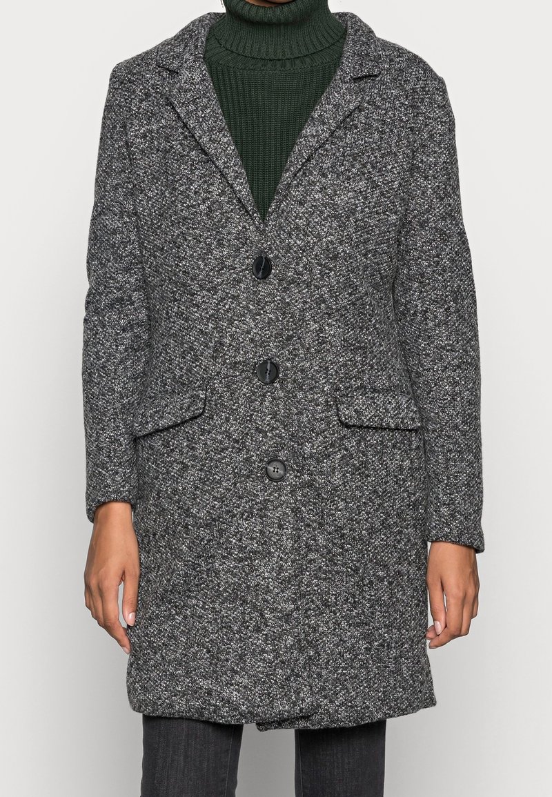 Femme portant un manteau gris texturé boutonné jusqu'aux genoux sur un pull à col roulé côtelé vert foncé.