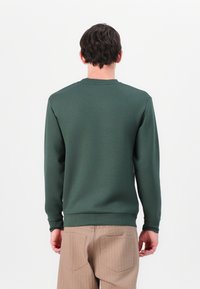Sweatshirt verde de gola redonda em tecido suave, com mangas longas e bainha canelada. Usado com calças de cordura bege.