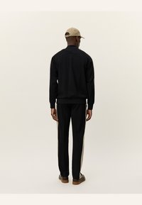 Black track jacket and pants with beige side stripes, made of smooth fabric. The outfit is paired with a beige cap and brown shoes.