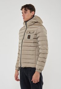 Giacca puffer beige con trapuntatura orizzontale, cappuccio, chiusura con zip e tasca sul petto con patch del logo. Maniche e orlo elasticizzati.